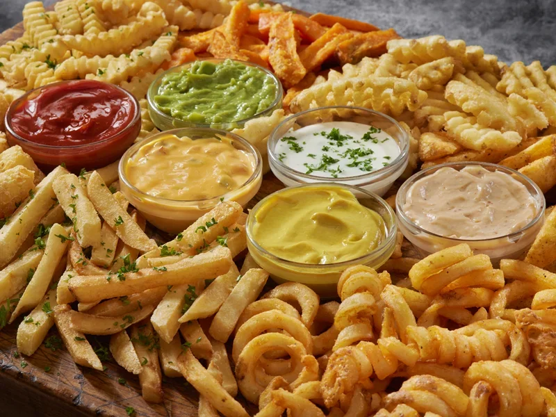 Assorted fried foods with dipping sauces including fries, onion rings, and waffle fries arranged on a wooden board.
