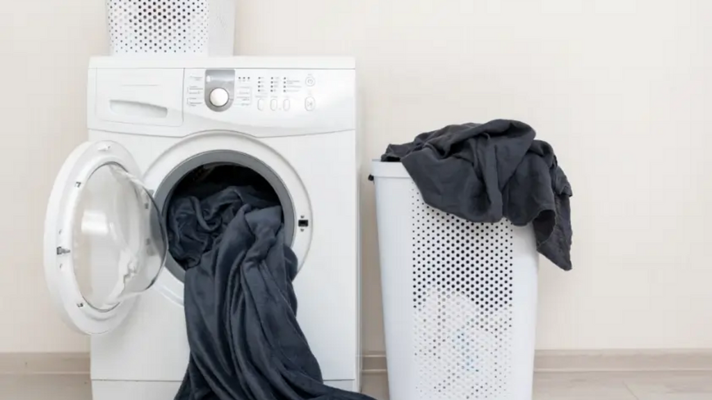 Máy giặt trắng với quần áo đen, giỏ giặt và giỏ để quần áo trên nền trắng.