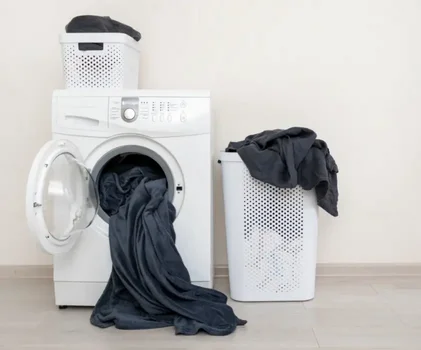Máy giặt trắng với quần áo đen, giỏ giặt và giỏ để quần áo trên nền trắng.