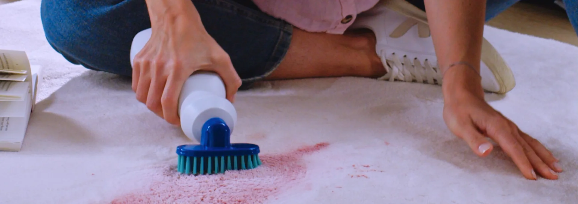 Person scrubbing a pink carpet stain with a blue brush and cleaning solution bottle.