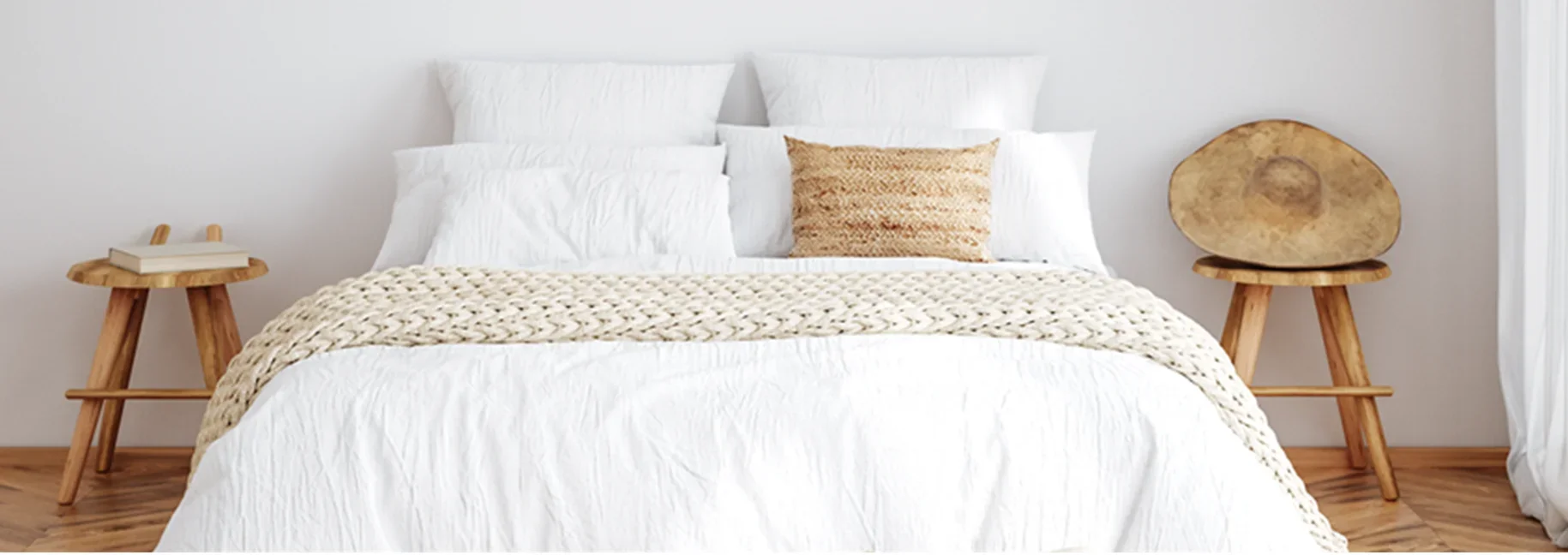 Modern bedroom with white bedding, woven beige throw blanket, wooden nightstands, and decorative woven hat accent.