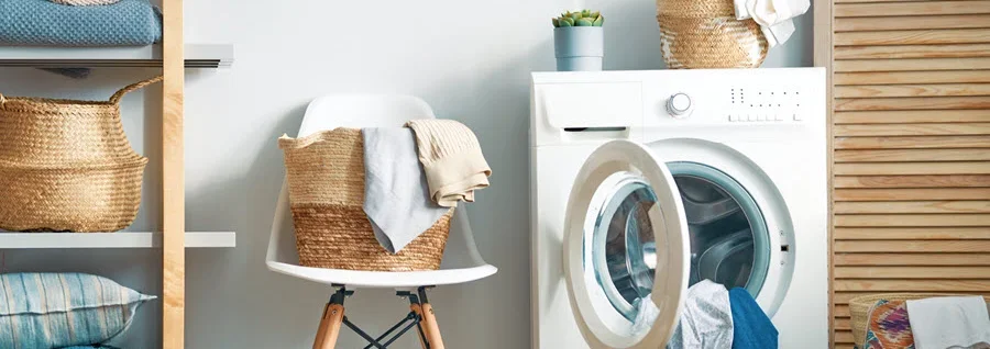 Organized laundry room with white washing machine, wooden shelving with folded linens and baskets, chair with towels, and striped flooring.