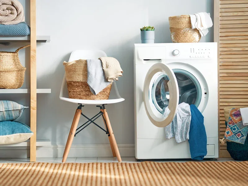 Organized laundry room with white washing machine, wooden shelving with folded linens and baskets, chair with towels, and striped flooring.