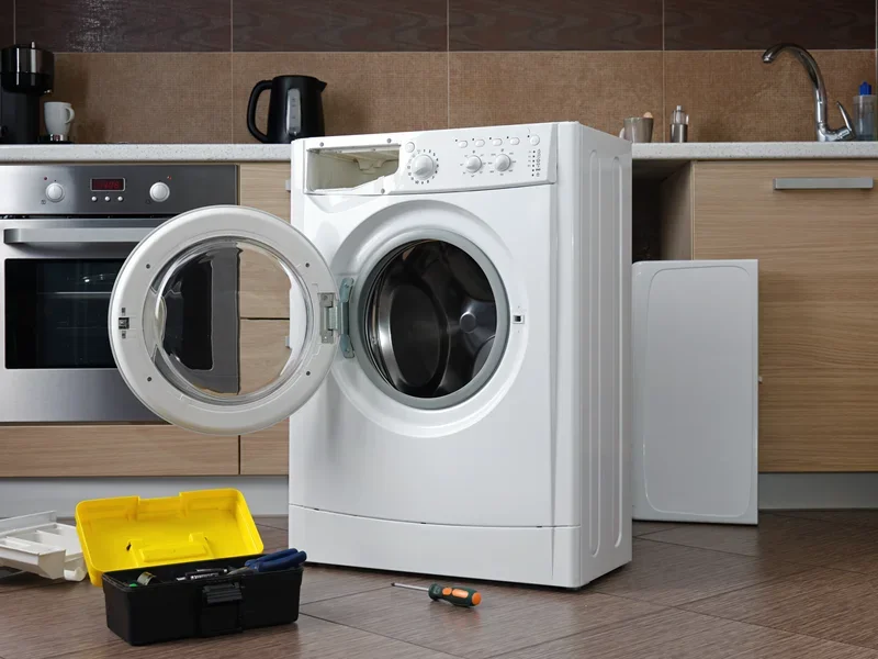 White front-load washing machine with open door in modern kitchen, surrounded by tools and appliances.
