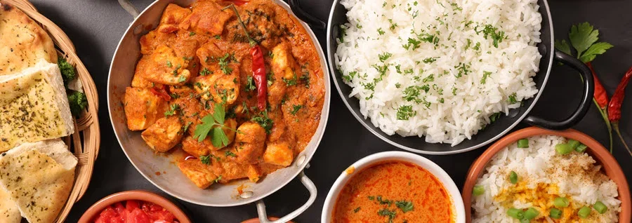 Overhead view of Indian cuisine spread featuring curry, rice, naan bread, and various colorful side dishes on dark background.