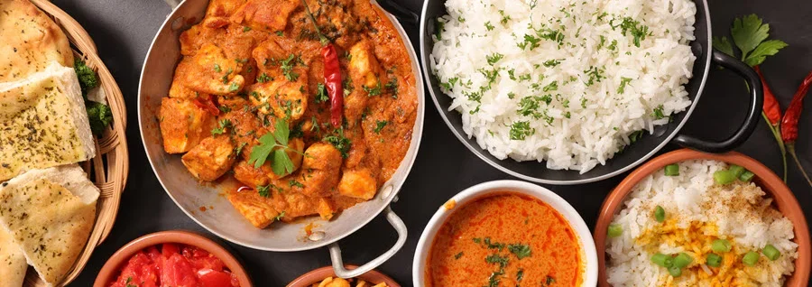 Overhead view of Indian cuisine spread featuring curry, rice, naan bread, and various colorful side dishes on dark background.