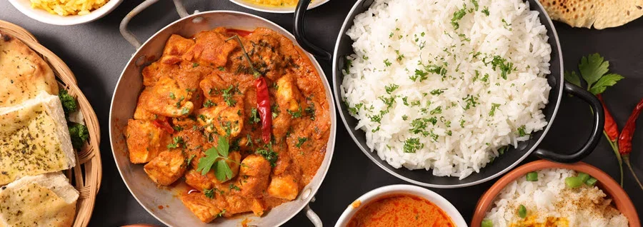 Overhead view of Indian cuisine spread featuring curry, rice, naan bread, and various colorful side dishes on dark background.