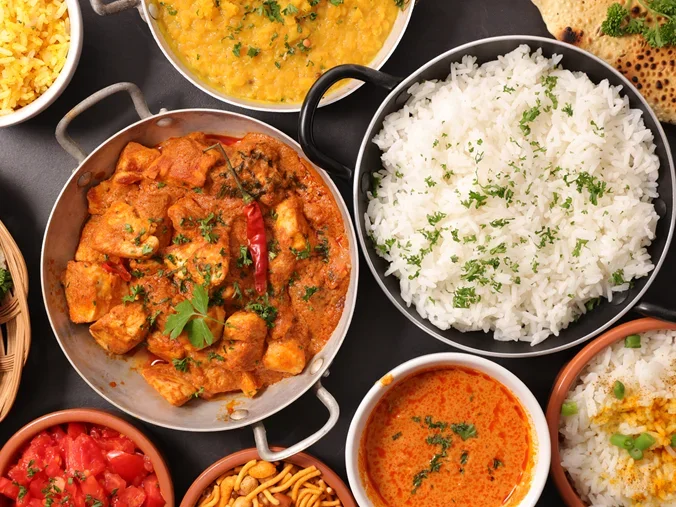 Overhead view of Indian cuisine spread with curry, rice, naan bread, and various side dishes on dark background.