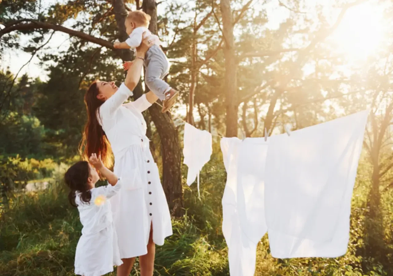 Một người mẹ nâng bé trai lên cao trong rừng, hai con gái mặc áo trắng đứng bên cạnh, quần áo trắng phơi trên dây.