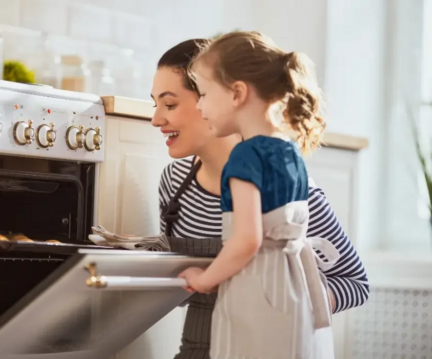 Một người phụ nữ và một bé gái đang mở lò nướng trong bếp hiện đại, cùng nhau chuẩn bị nấu ăn.