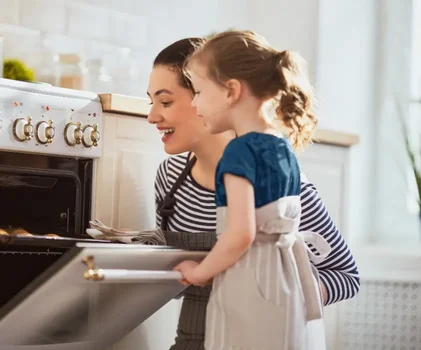 Một người phụ nữ và một bé gái đang mở lò nướng trong bếp hiện đại, cùng nhau chuẩn bị nấu ăn.