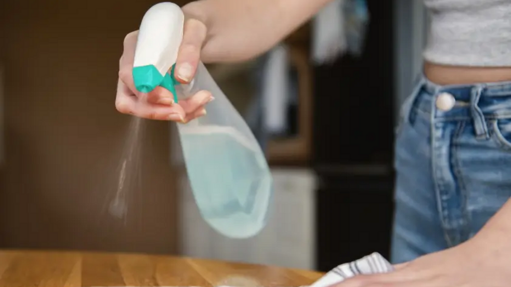 Một người đang phun chất lỏng từ một chai xịt có nắp xanh lên một miếng vải trắng trên bàn gỗ.