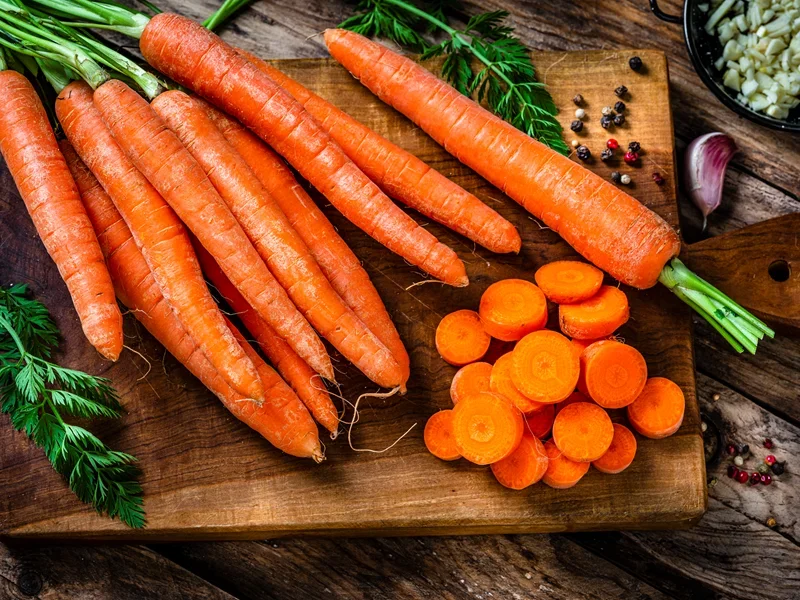 Fresh carrots with green tops and sliced rounds on a wooden cutting board, surrounded by herbs and spices.