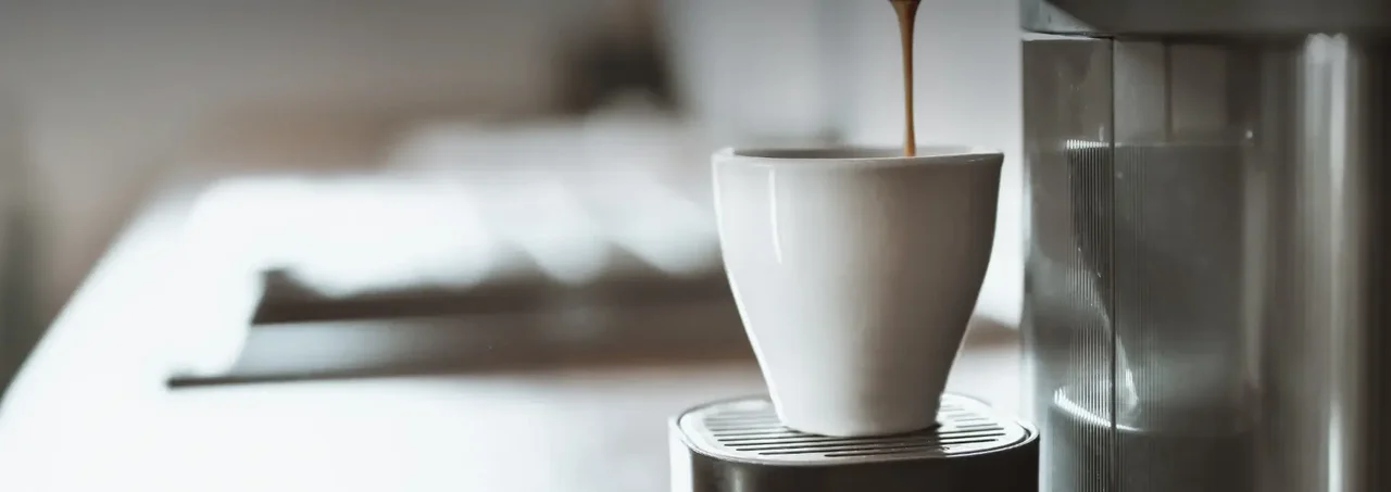 Cà phê màu nâu được đổ vào cốc trắng trên máy pha cà phê, với phông nền mờ của văn phòng hiện đại.