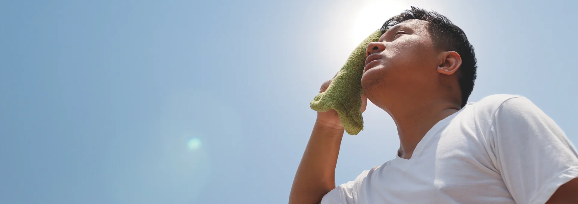 Man in white shirt wiping sweat from his face with a green towel against a bright blue sky.