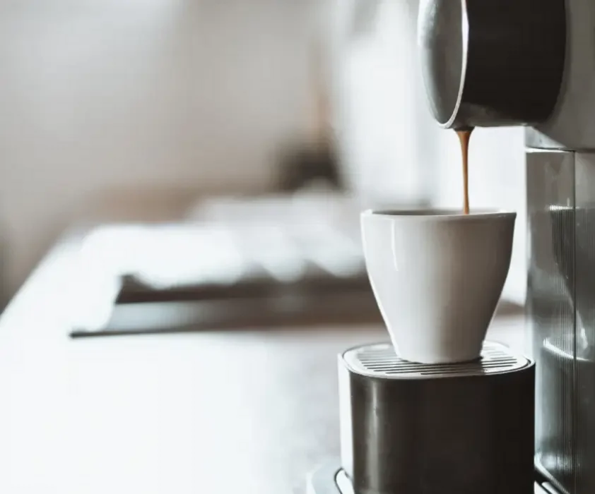 Máy pha cà phê đang rót cà phê nâu vào tách trắng trên mặt bàn trắng sáng.