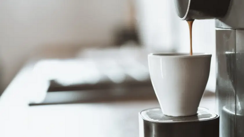 Máy pha cà phê đang rót cà phê nâu vào tách trắng trên mặt bàn trắng sáng.