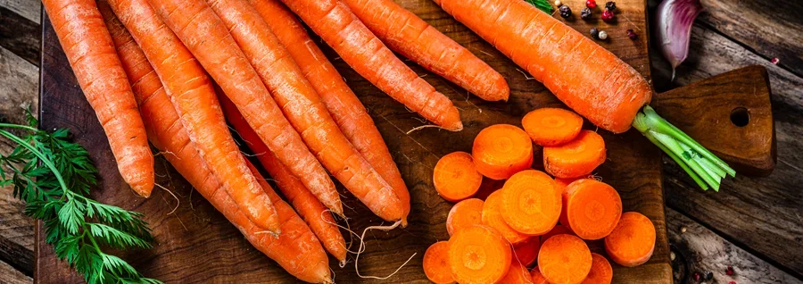 Fresh carrots with green tops and sliced carrot rounds on a wooden cutting board surrounded by herbs and spices.