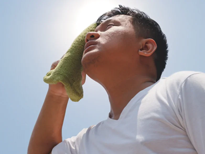 Man wiping sweat from his face with a green towel on a sunny day, looking upward.