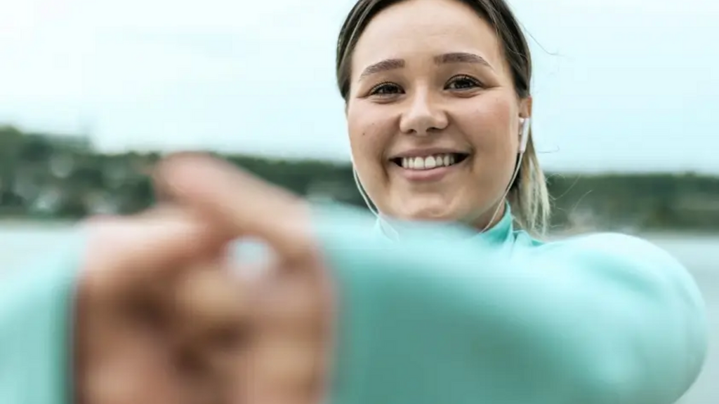 一位年轻女性穿着浅绿色上衣，在户外水边微笑并伸出手指向镜头，背景是模糊的河流和树木。